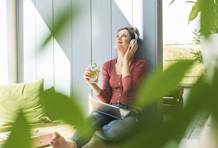 © Westend61/Westend61 via Getty Images Lächelnde Frau mit Kopfhörern und Laptop, die zu Hause am Fenster sitzt  | © Westend61/Westend61 via Getty Images