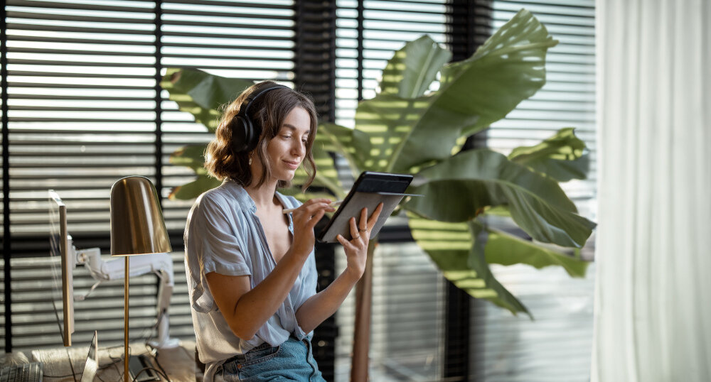 Junge Frau arbeitet an einem Touchpad und fühlt sich wohl in ihrem gemütlichen Heimbüro mit einer Grünpflanze im Hintergrund | © RossHelen/Shutterstock.com