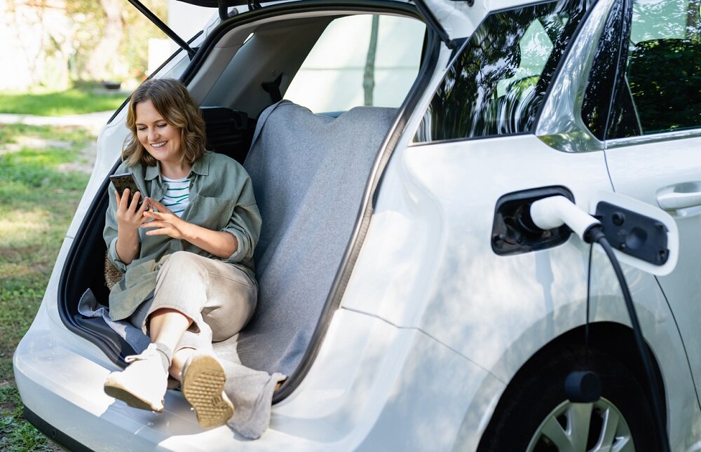 Elektromobilitaet bei ALDI Grüne Energie | © © iStock.com | Scharfsinn86 Eine Frau sitzt im geöffneten Kofferraum ihres Elektroautos und lächelt freudig auf ihr Smartphone-Display, während das Auto geladen wird. | © © iStock.com | Scharfsinn86