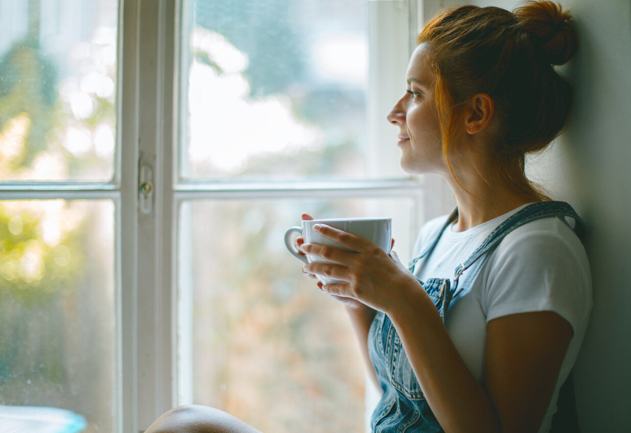 Rothaarige Frau mit Latzhose hält Kaffeetasse und schaut aus dem Fenster. | © iStock-1047712036